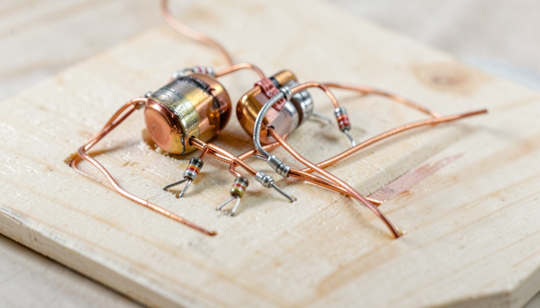 A close-up of a hand-built passive crossover on a plywood board, with large foil inductors, precision resistors, and neatly zip-tied wiring in warm copper and silver tones against pale wood grain.